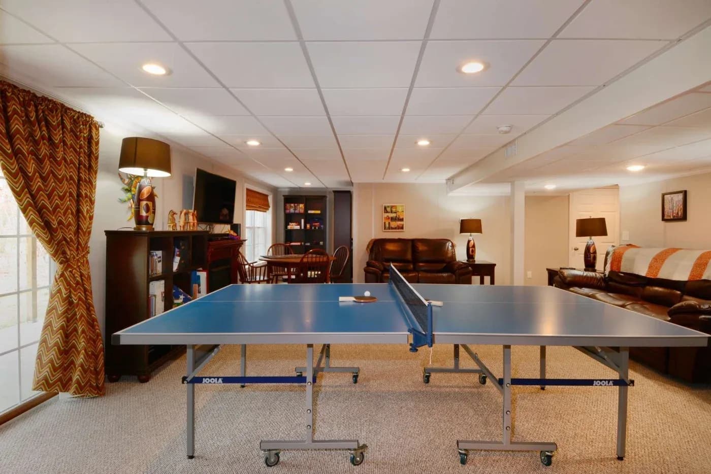 A spacious basement features a blue ping pong table centered on beige carpet. Surrounding it are cozy leather couches, a bookshelf, and warm lighting.
