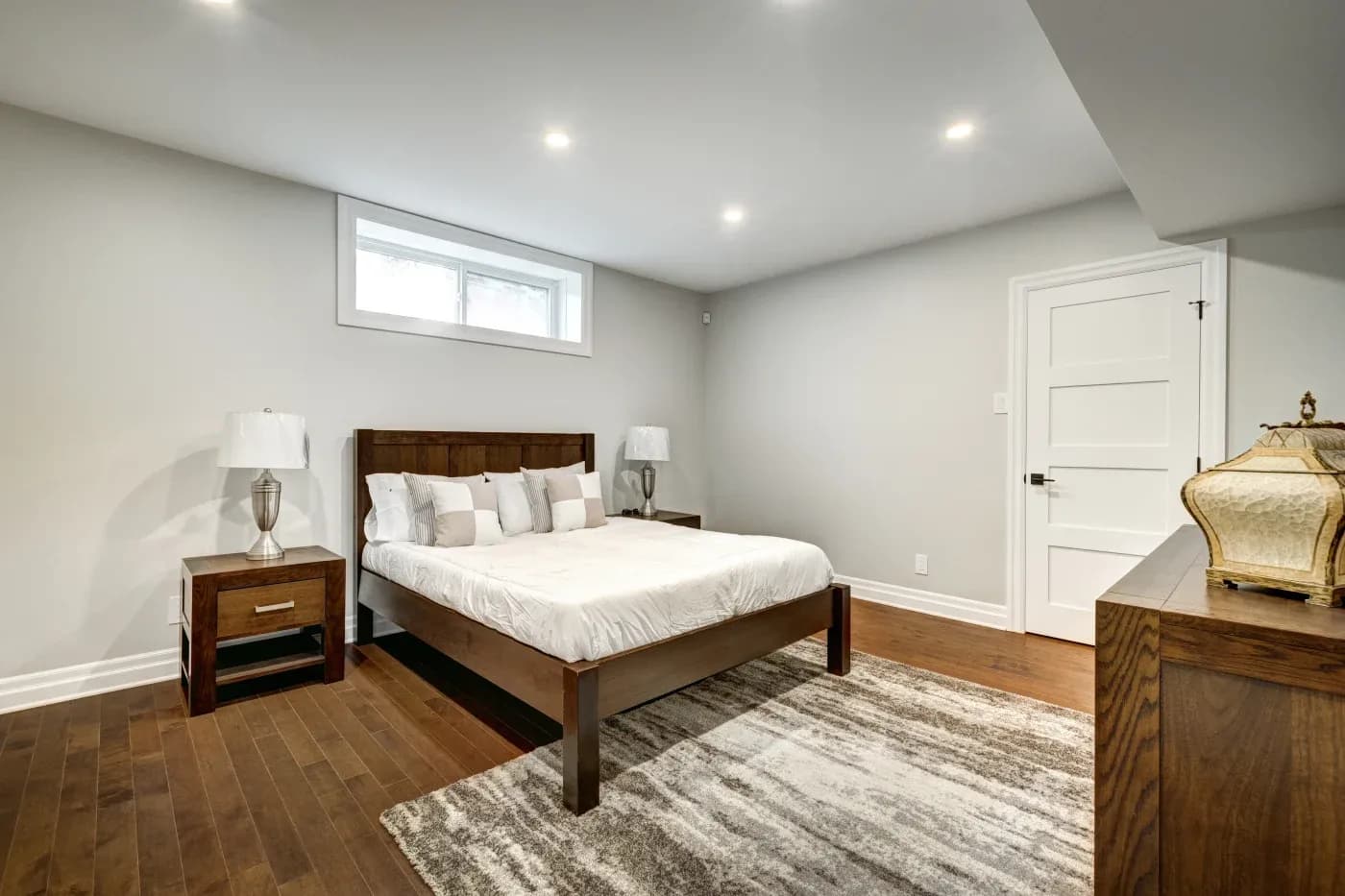 A modern bedroom with a wooden bed, white bedding, and matching side tables with lamps. A soft, patterned rug lies on the hardwood floor. The room feels serene and minimalistic.