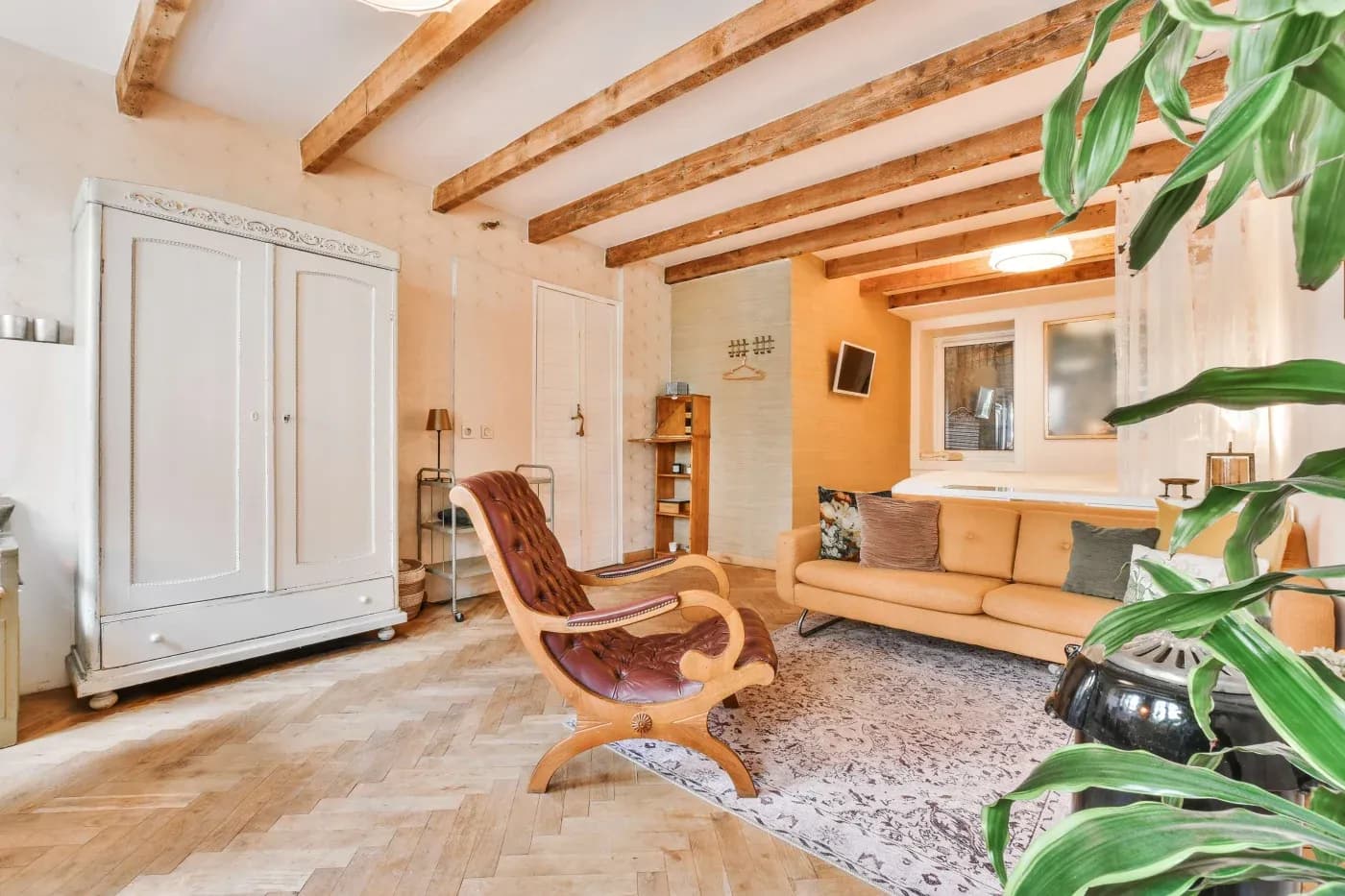 Cozy living room with wooden beams, a mustard sofa, and a brown lounge chair. A white wardrobe and green plant add warmth and style.