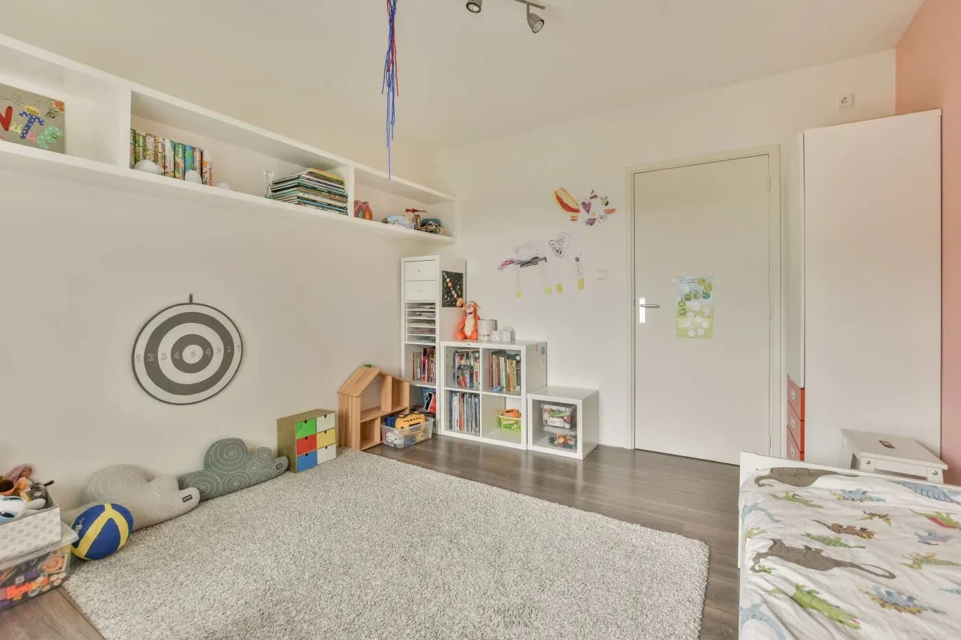Bright children's room with a plush rug, shelves filled with books and toys, playful wall decor, a dartboard, and a bed with animal-themed bedding.