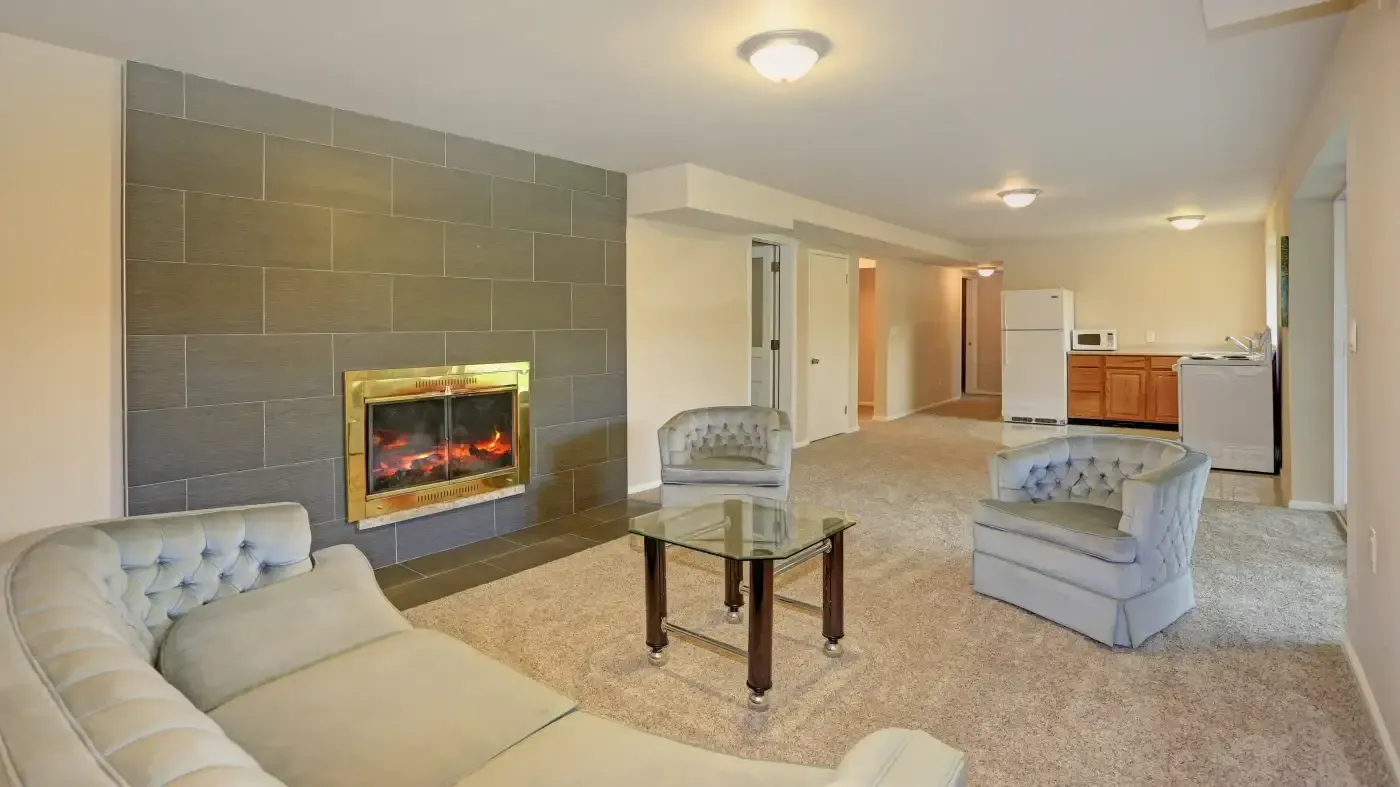 A cozy living room features a gray sectional sofa and armchairs around a glass coffee table. A lit fireplace on a tiled wall adds warmth. A small kitchen area is visible in the background, contributing to a homely atmosphere.