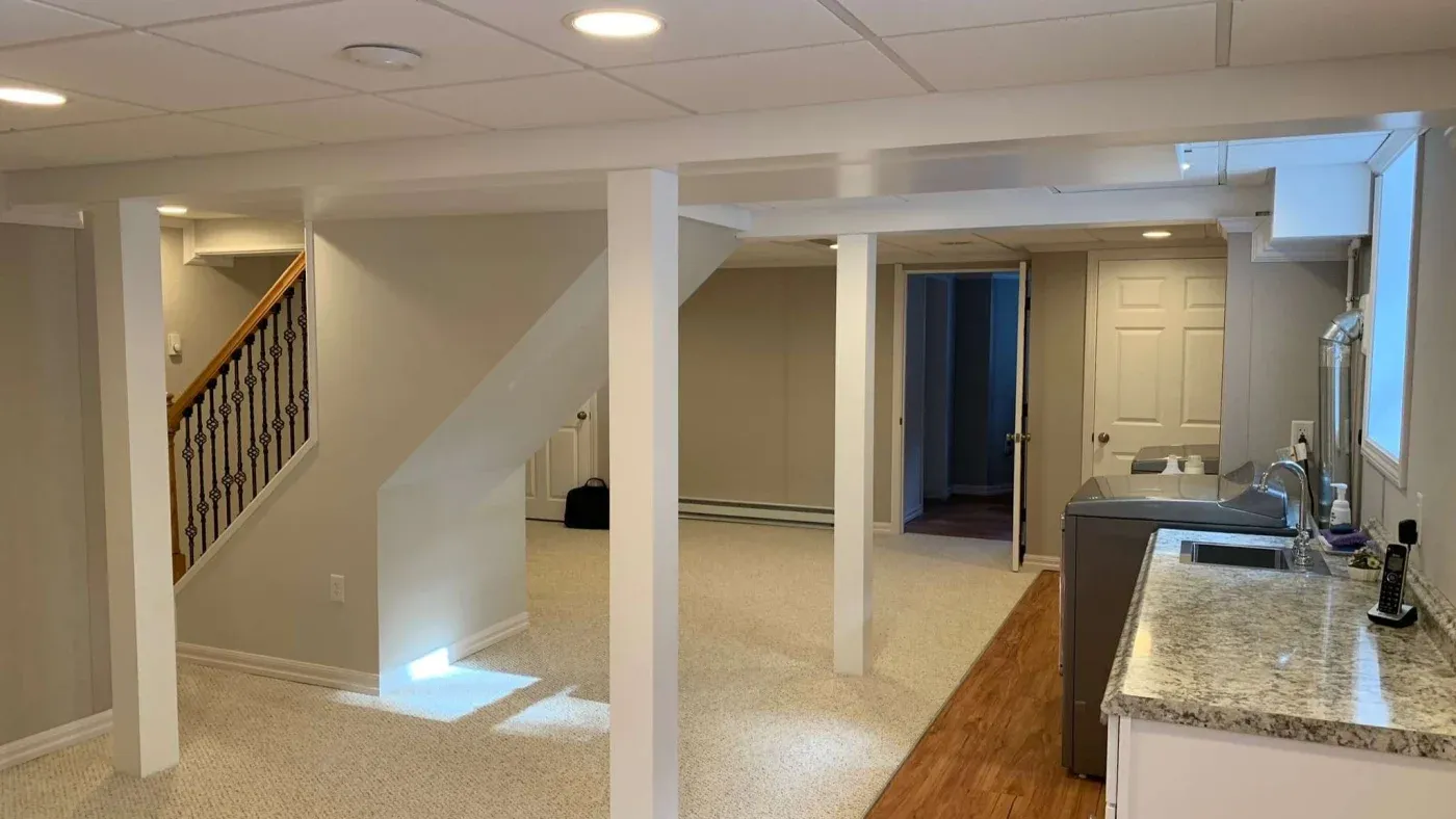 Finished basement with beige walls and carpet, featuring a staircase, columns, and recessed lighting. A functional area with a counter, sink, and washer-dryer.
