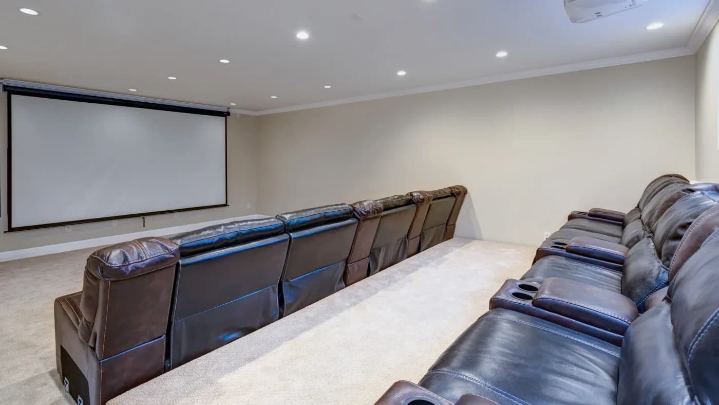 Spacious home theater with plush reclining seats and cup holders facing a large, blank projection screen. The room has a cozy, inviting ambiance.