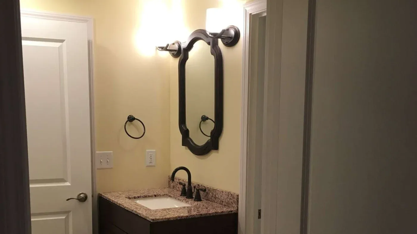 A small bathroom with a wooden vanity and granite countertop, ornate mirror, and bright lighting. Neutral tones create a clean, warm atmosphere.