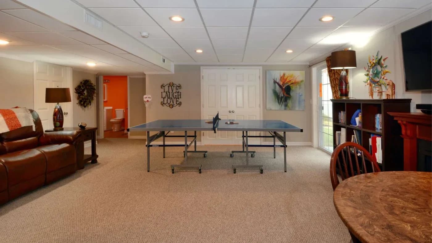 Basement game room with a ping pong table centered on beige carpet. Cozy seating area with a leather sofa, warm lighting, and vibrant wall art.