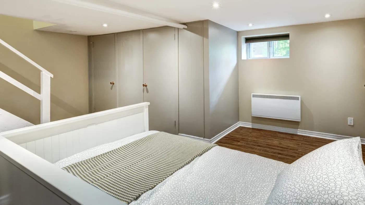 Small, minimalist bedroom with neutral tones. Features a bed with patterned bedding, a narrow window, a radiator, and a stairway to the left. Cozy ambiance.