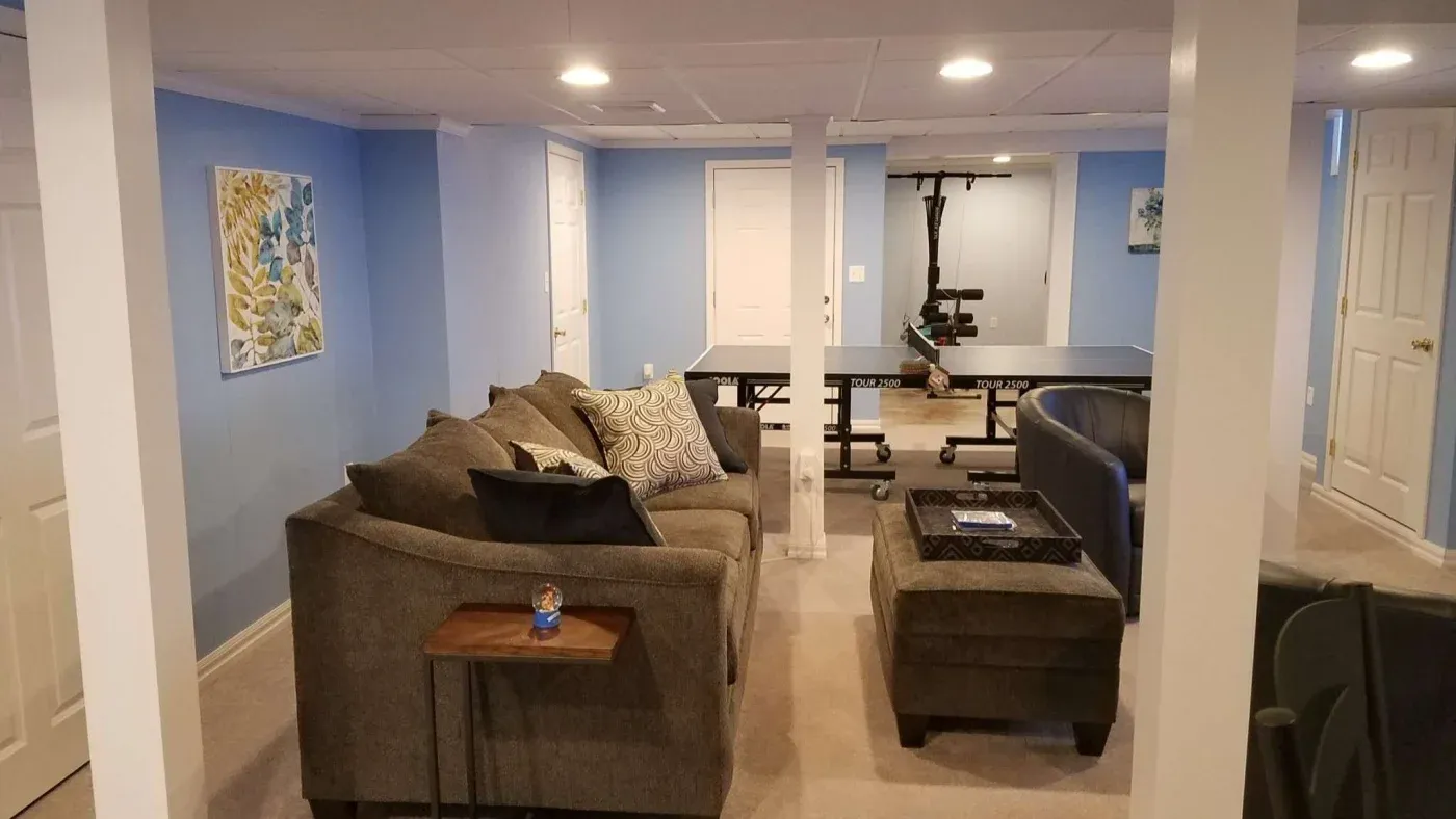 Basement recreation room with a cozy gray sectional sofa, accent pillows, and a wooden side table. A ping pong table and blue walls create a relaxed atmosphere.