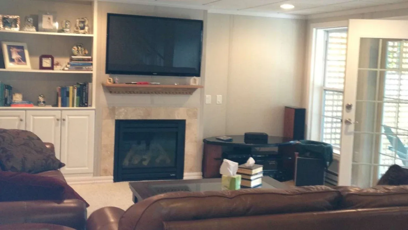 A cozy living room with a fireplace and TV mounted above. Shelves with books and photos flank the fireplace. Large windows and a glass door provide natural light. Brown leather couches add warmth.