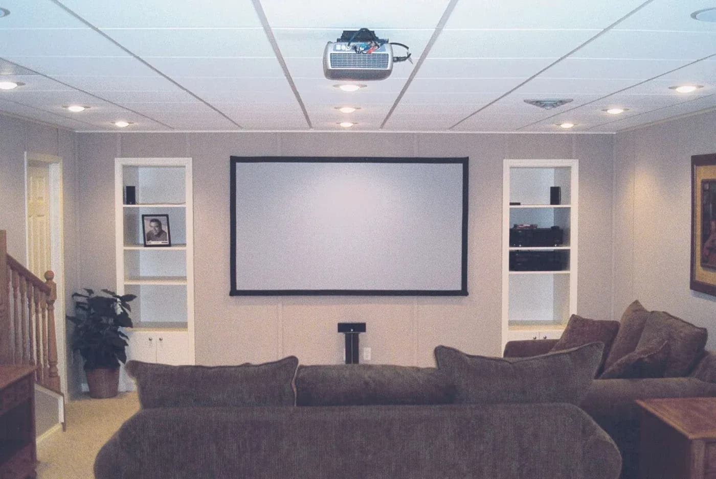 A cozy home theater with a large screen, brown couches, shelves with decor and electronics, a ceiling projector, and soft lighting sets a relaxing mood.
