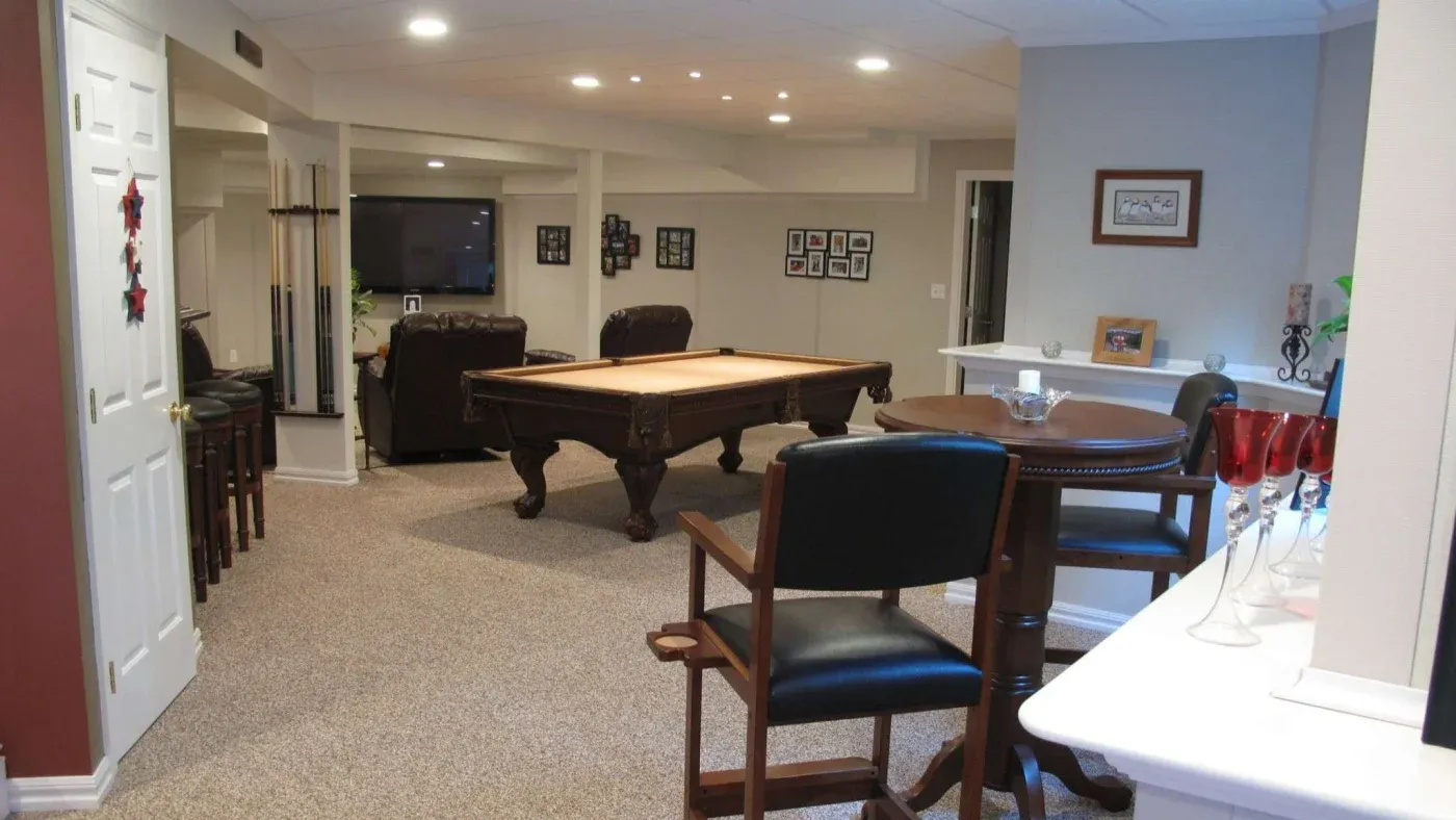 A cozy basement game room features a central pool table, surrounded by leather chairs and bar stools. Soft lighting and photo frames create a warm, inviting atmosphere.