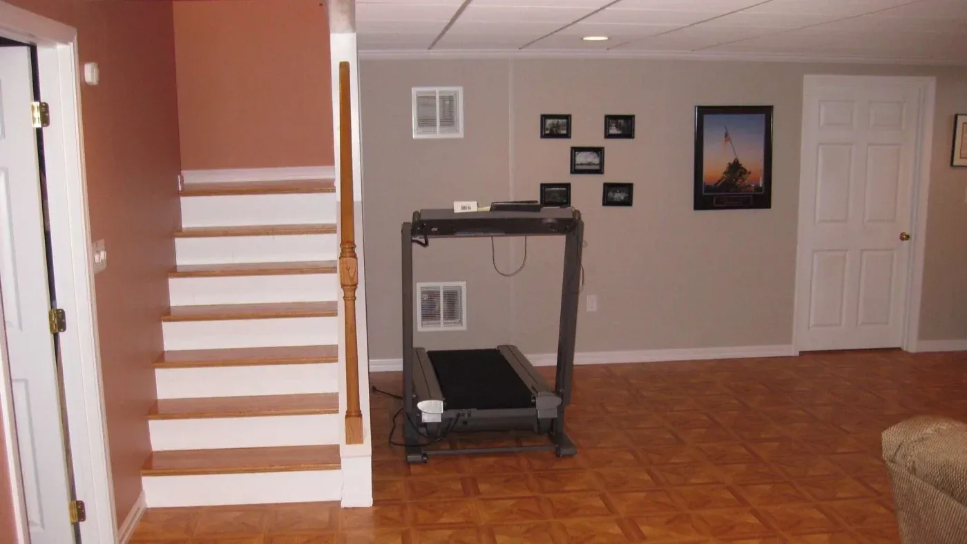 A basement room with wood-patterned flooring, a treadmill against the wall, several small framed photos, and white stairs leading up, conveys a calm, organized space.