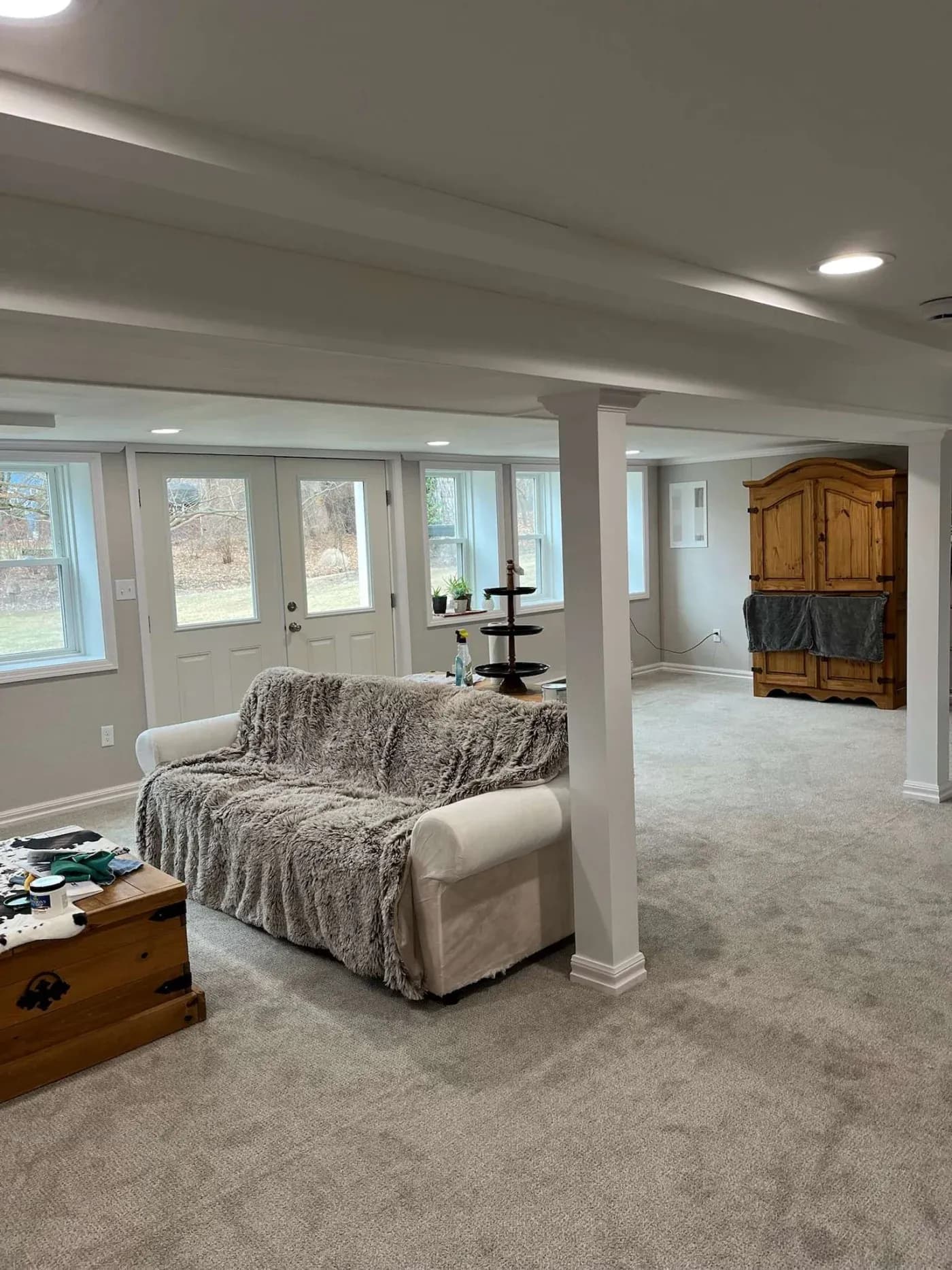 Spacious, well-lit basement with beige carpet, white sofa draped with a gray blanket, wooden armoire, and windows illuminating the cozy room.