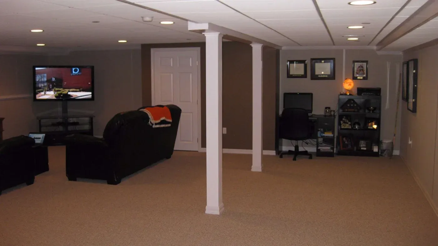 A cozy basement features a TV area with dark sofas, a home office with a desk and computer, and framed pictures on taupe walls, conveying a relaxed ambiance.
