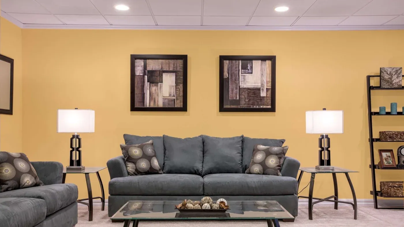 A cozy living room with a yellow wall, featuring a gray sofa and armchair adorned with decorative pillows. Two abstract paintings hang above the sofa, flanked by lit table lamps. A glass coffee table displays decorative items, adding a modern touch.