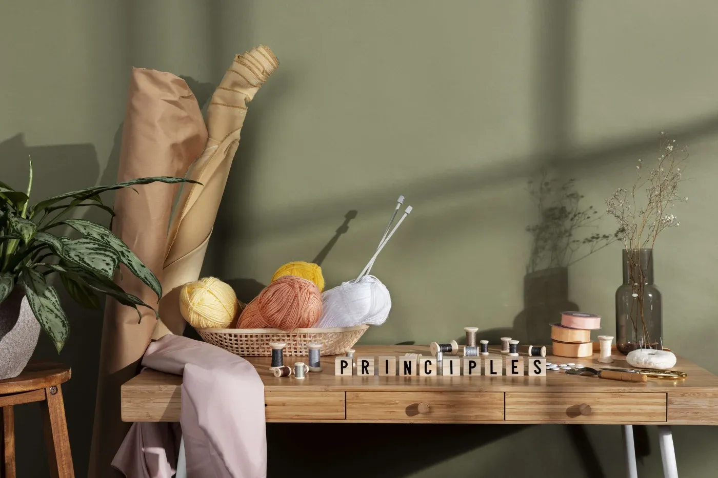 A wooden table with sewing supplies, yarn, and knitting needles. The word "PRINCIPLES" is spelled out in blocks. A potted plant and fabric rolls are nearby.