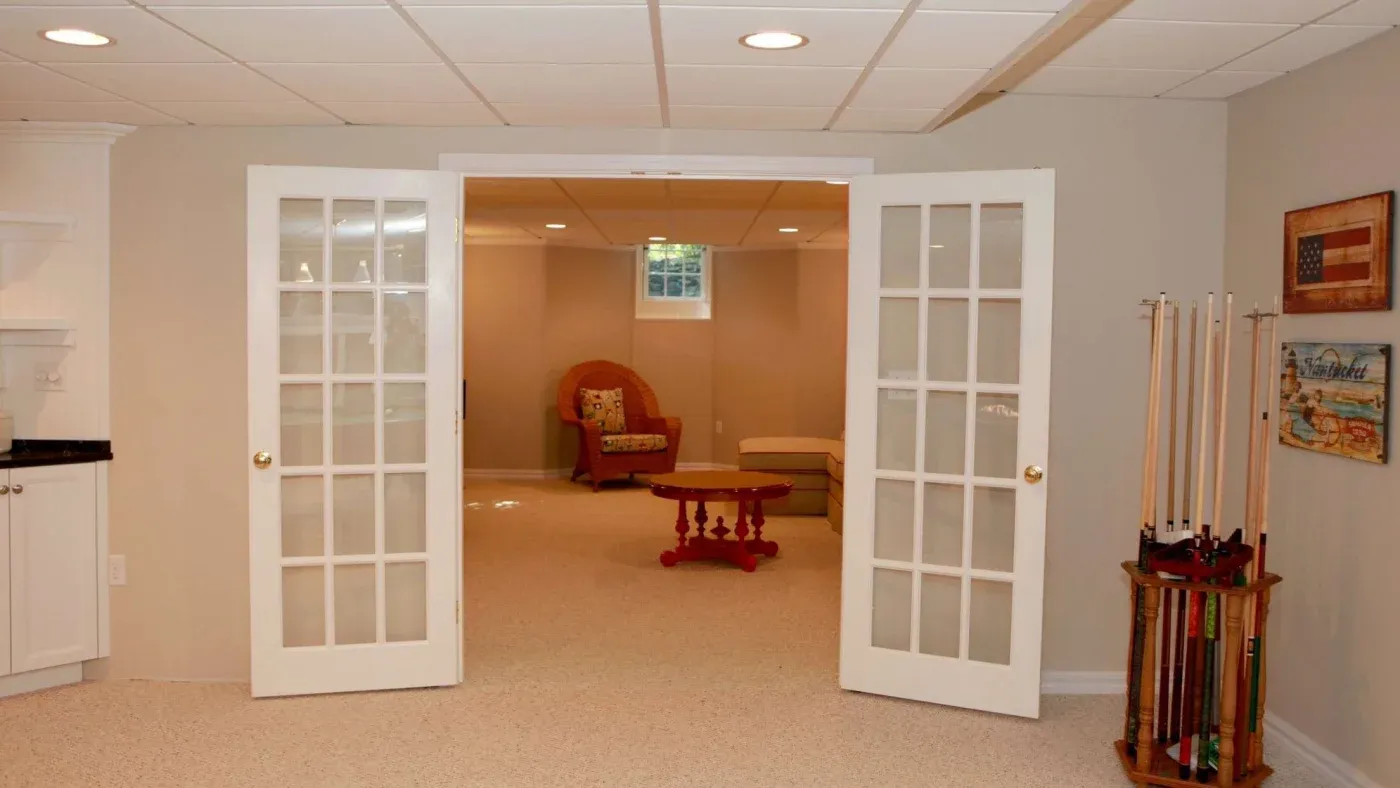 Basement room with open French doors, armchair, red round table, pool cues, and wall art. Bright, cozy atmosphere with beige walls and carpet.