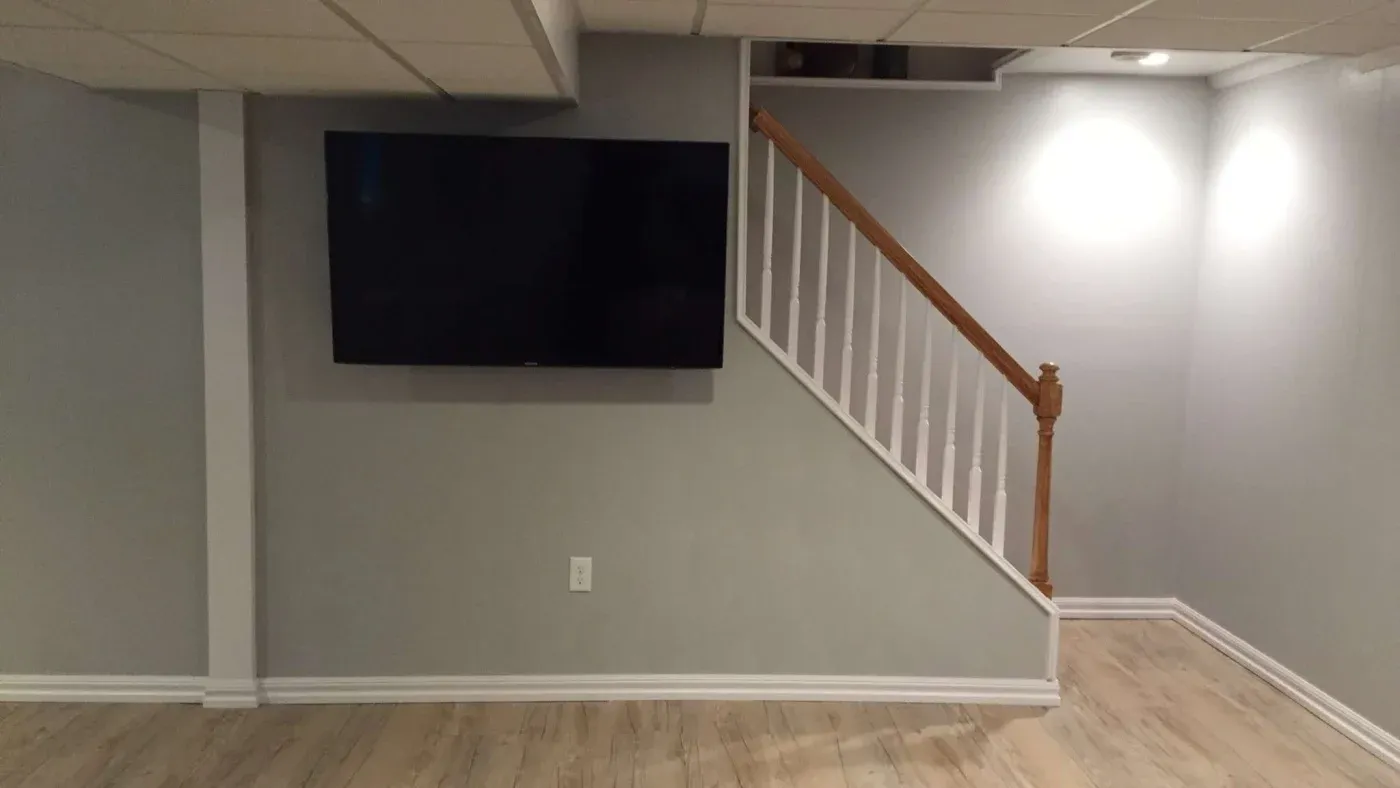A modern basement room with light gray walls and wood flooring features a large flat-screen TV mounted on the wall. A staircase with a wooden handrail ascends to the right, leading to a brightly lit upper area. The space feels clean and minimalistic.