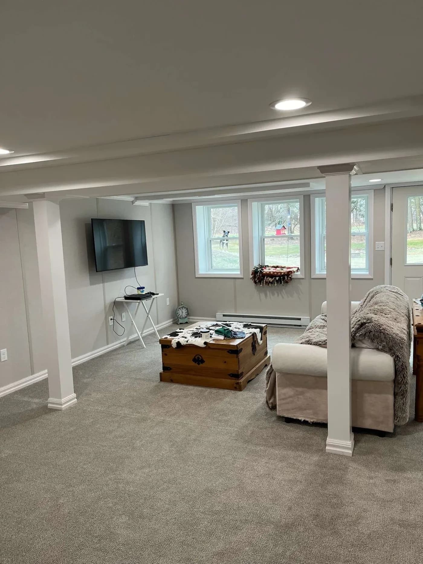 Cozy basement room with beige carpet, a wall-mounted TV, and a light brown sofa. A wooden chest serves as a coffee table near large windows.