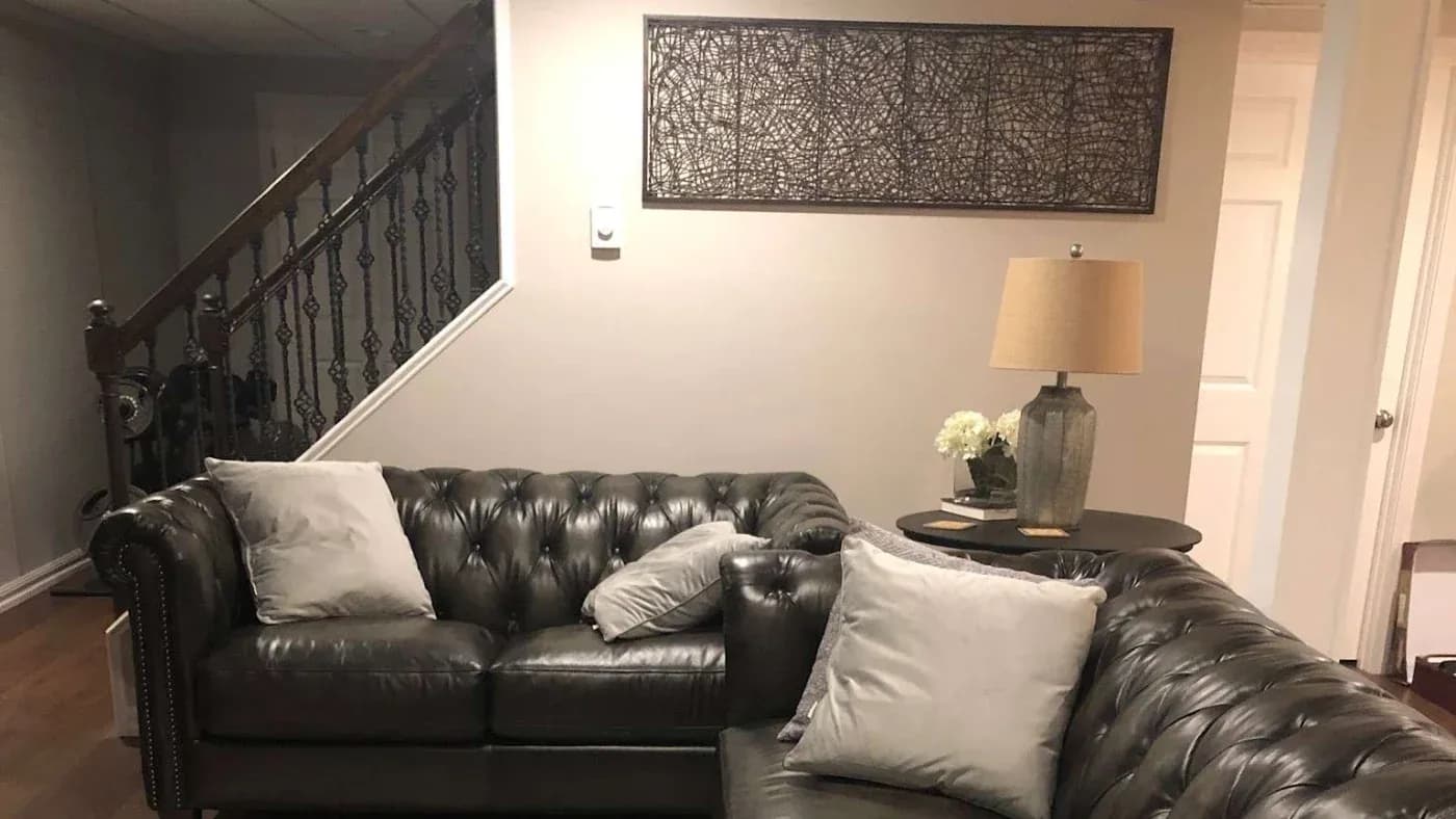 A cozy living room with dark leather sofas adorned with light gray cushions. A warm lamp and floral decor sit on a side table. An abstract wall art piece hangs behind them, next to a staircase with ornate railings, conveying a welcoming and stylish atmosphere.
