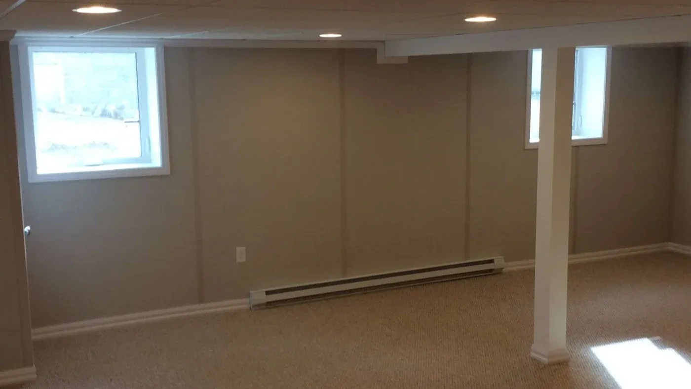 A dimly-lit, empty basement room with beige carpet, cream walls, and two small windows. Recessed lights create soft illumination, conveying a calm atmosphere.