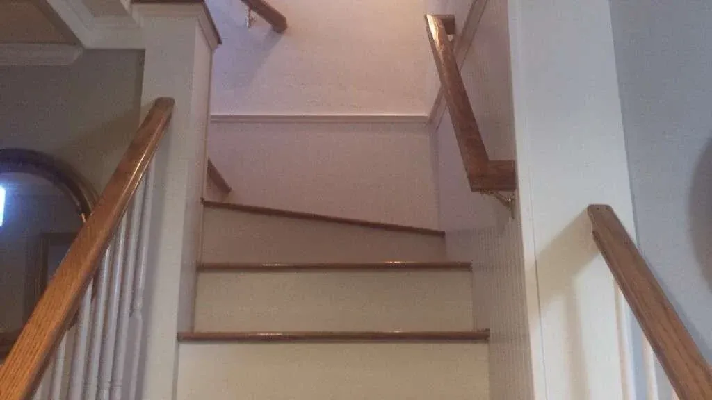A flight of wooden stairs with white risers leads up to a landing. The handrail curves to the right. The lighting is warm and natural.