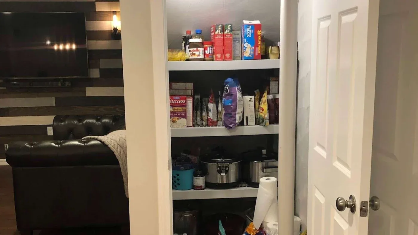 A small pantry with white shelves stocked with food items, snacks, and cooking supplies. Nearby, a living room features a black couch and a wall-mounted TV.