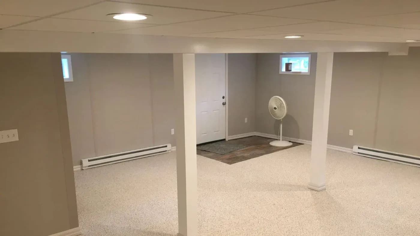 Spacious, empty basement with beige carpet, light gray walls, and white ceiling. A closed white door and an oscillating fan are seen, creating a calm, neutral atmosphere.