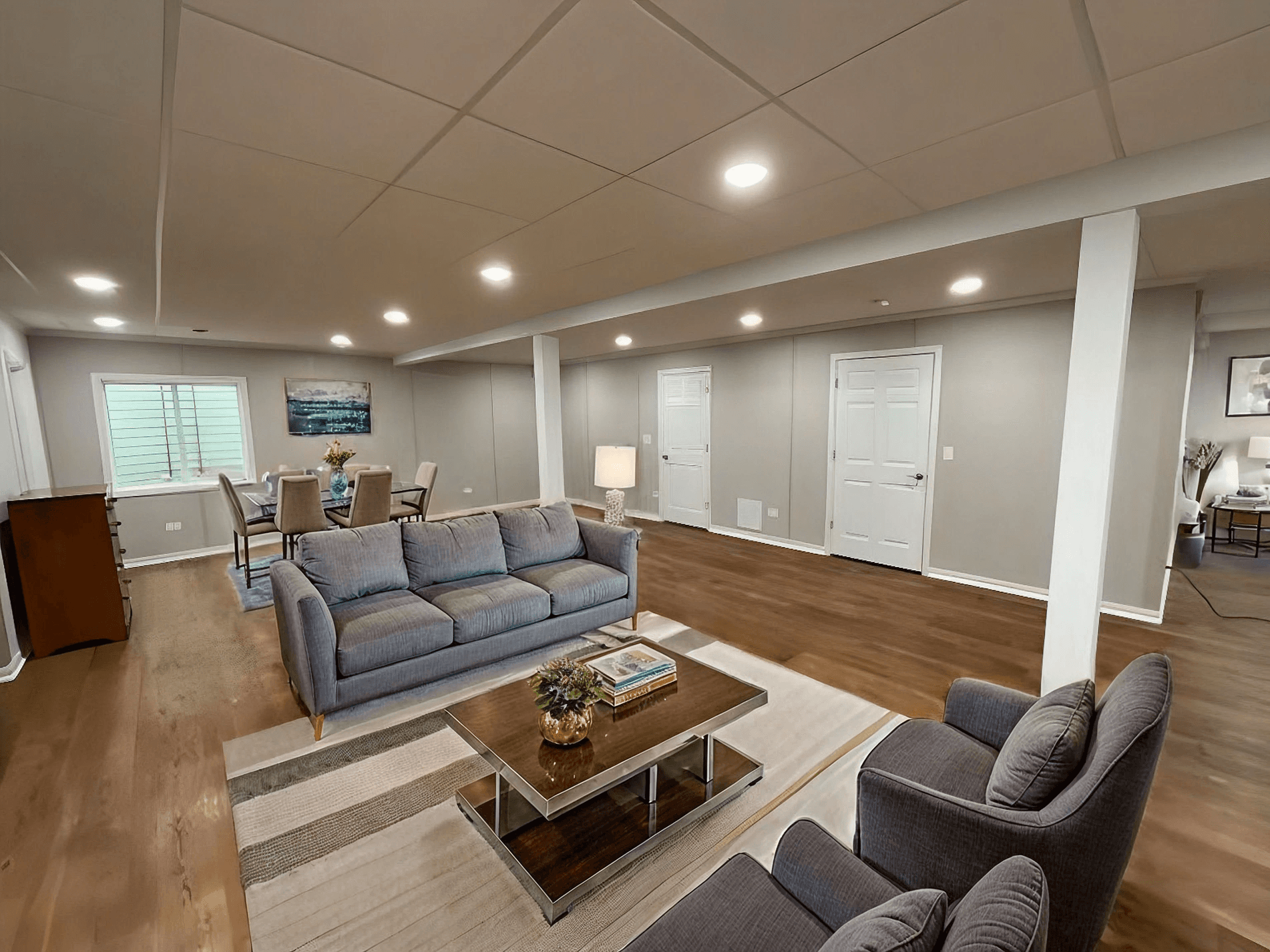 Spacious basement with modern gray sofas, armchairs, and a wooden coffee table on a striped rug. Soft lighting, neutral tones, and dining area in background.