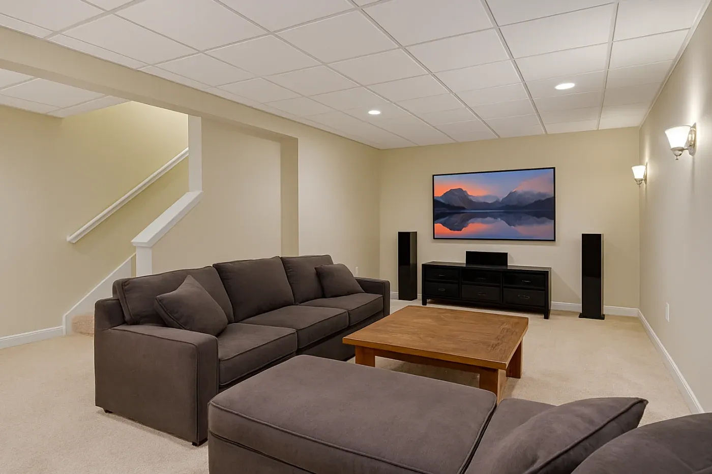 Modern basement with beige walls, featuring a large gray sectional sofa and wooden coffee table. A flat-screen TV displays a serene landscape.