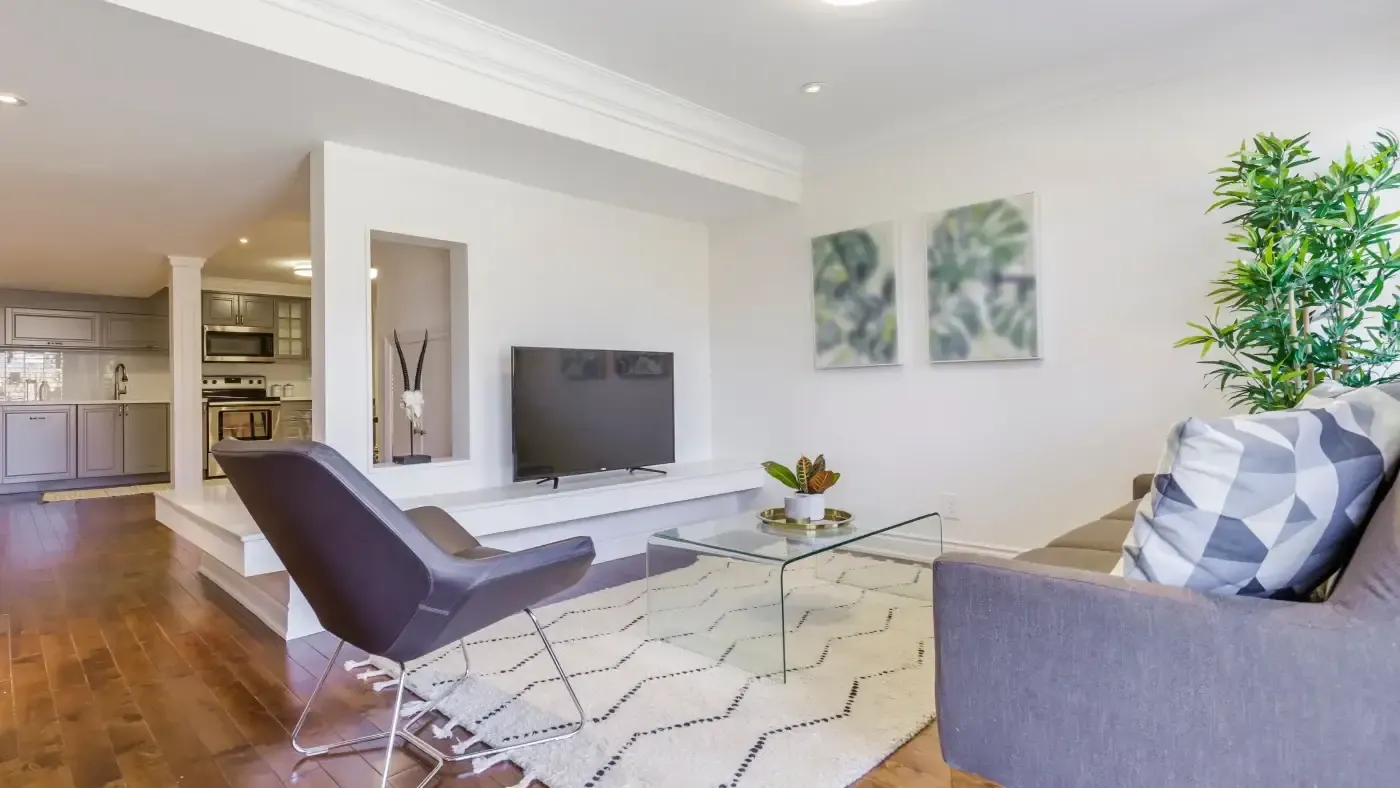 Modern living room with a sleek gray sofa, geometric cushions, and a glass coffee table on a patterned rug. A TV is mounted on a shelf, and a plant adds greenery. The open kitchen is visible in the background. Bright and inviting ambiance.
