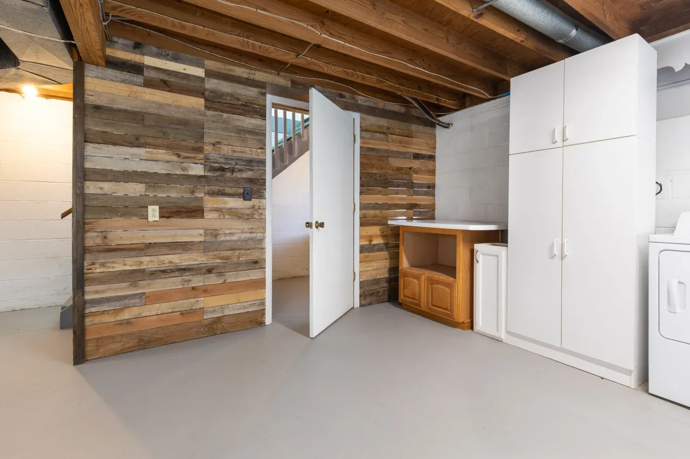 Basement with rustic wood accent wall, open white door, exposed beams, white cabinets, and washing machine. Cozy and functional space.