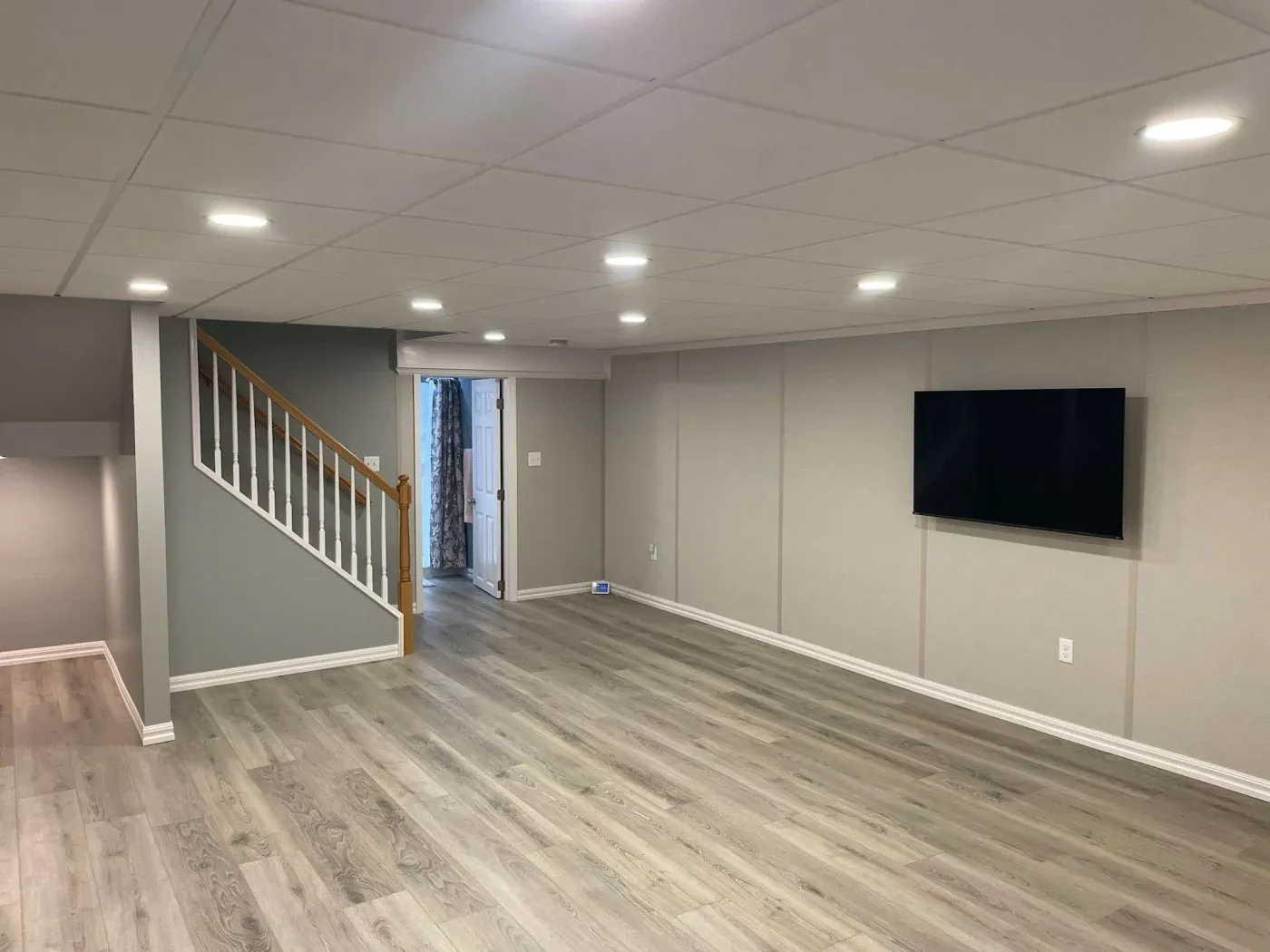 A modern basement with light wood flooring and gray walls, illuminated by recessed ceiling lights. A flat-screen TV is mounted on the right wall, and stairs with a wooden handrail lead up on the left. A door at the rear opens to another room. The space feels open and inviting.