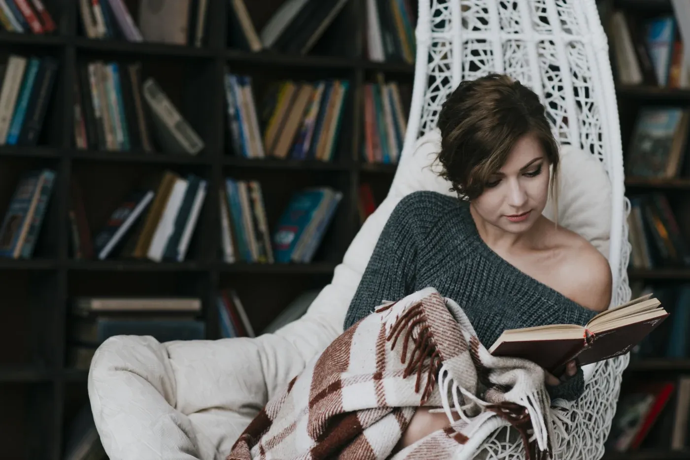 A woman in a cozy gray sweater reads a book while sitting in a white hanging chair. She is wrapped in a plaid blanket, with a bookshelf in the background.