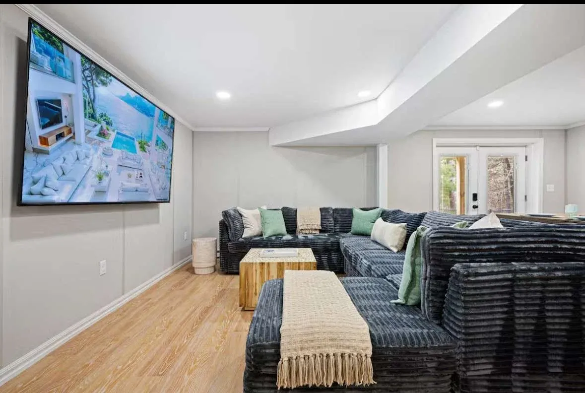 Spacious living room with a large sectional sofa, soft pillows, and a textured throw. A big-screen TV displays a scenic view. Bright, cozy ambiance.