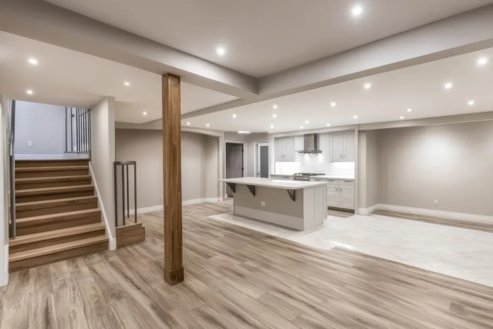 Modern basement with light wood flooring and recessed lighting. Features include a staircase, wooden column, and kitchen with an island. Spacious and minimalist.