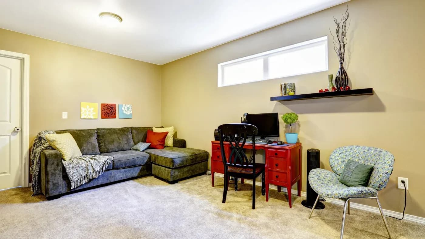 Cozy room with a gray sectional sofa topped with a throw blanket and bright pillows. A vibrant red desk holds a computer. Modern art and decor add charm.