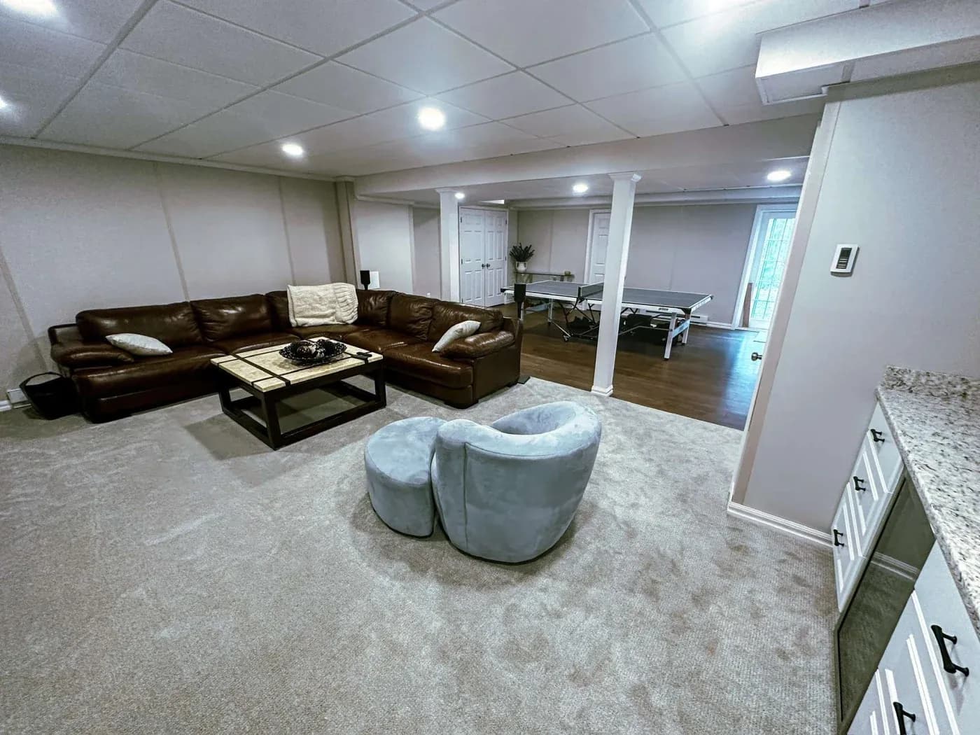 Spacious basement with light carpet, featuring a large brown leather sectional sofa, a coffee table, and two blue chairs. A ping pong table is visible in the background.