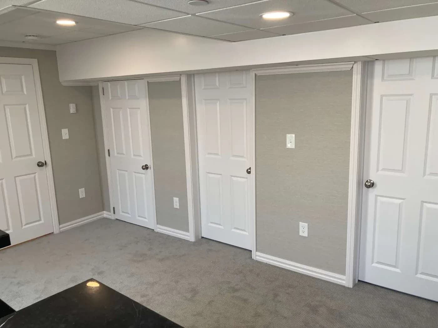 A room with four closed white doors on a light gray wall. Gray carpet covers the floor, and recessed ceiling lights create a modern, clean look.