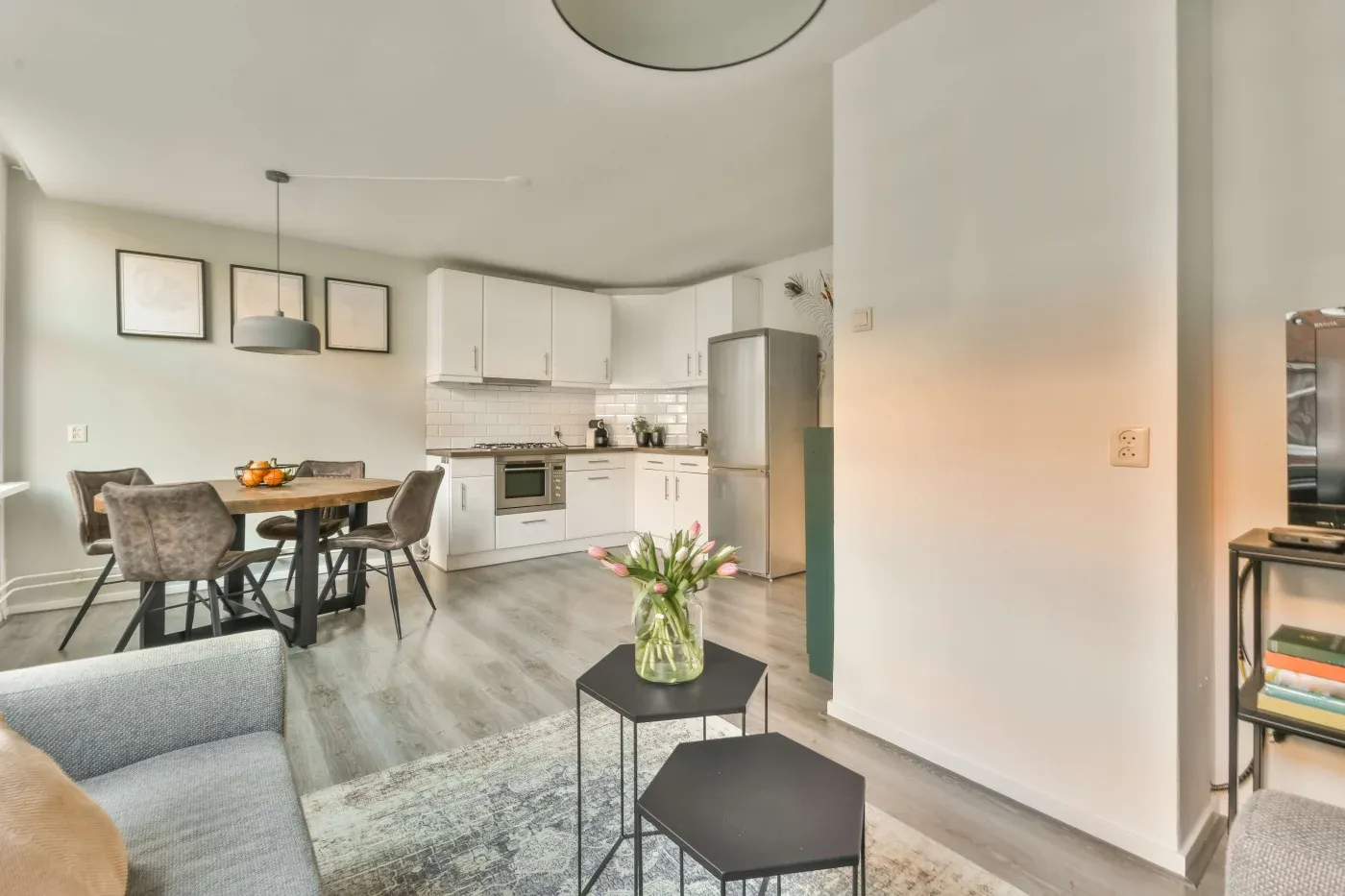 Modern open-plan living area with a cozy kitchen in white tones. A dining table with chairs is on the left, tulips on a coffee table center the room.