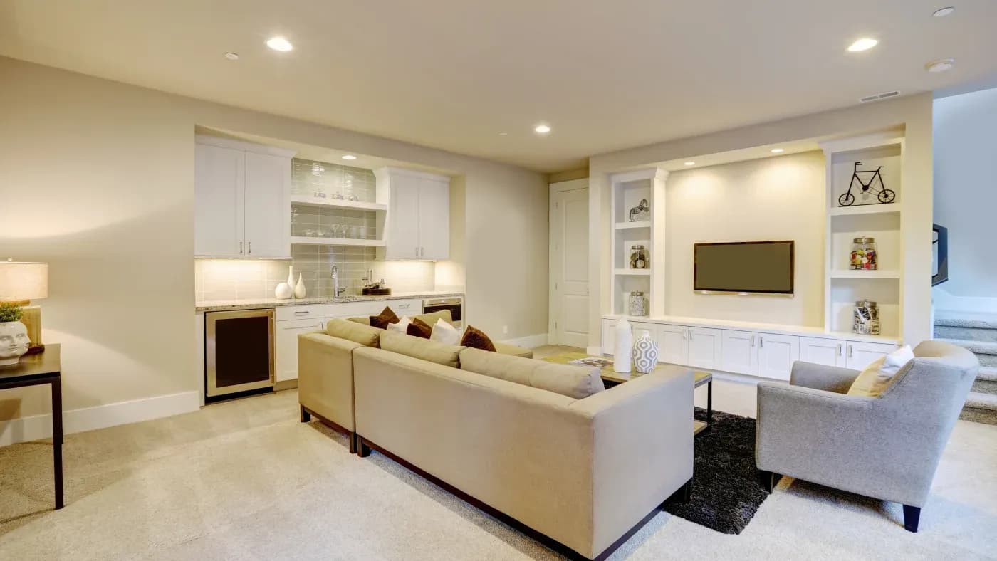 Spacious living room with beige sofa, armchair, and a flat-screen TV on a wall unit. Warm lighting and a cozy atmosphere with a kitchenette nearby.