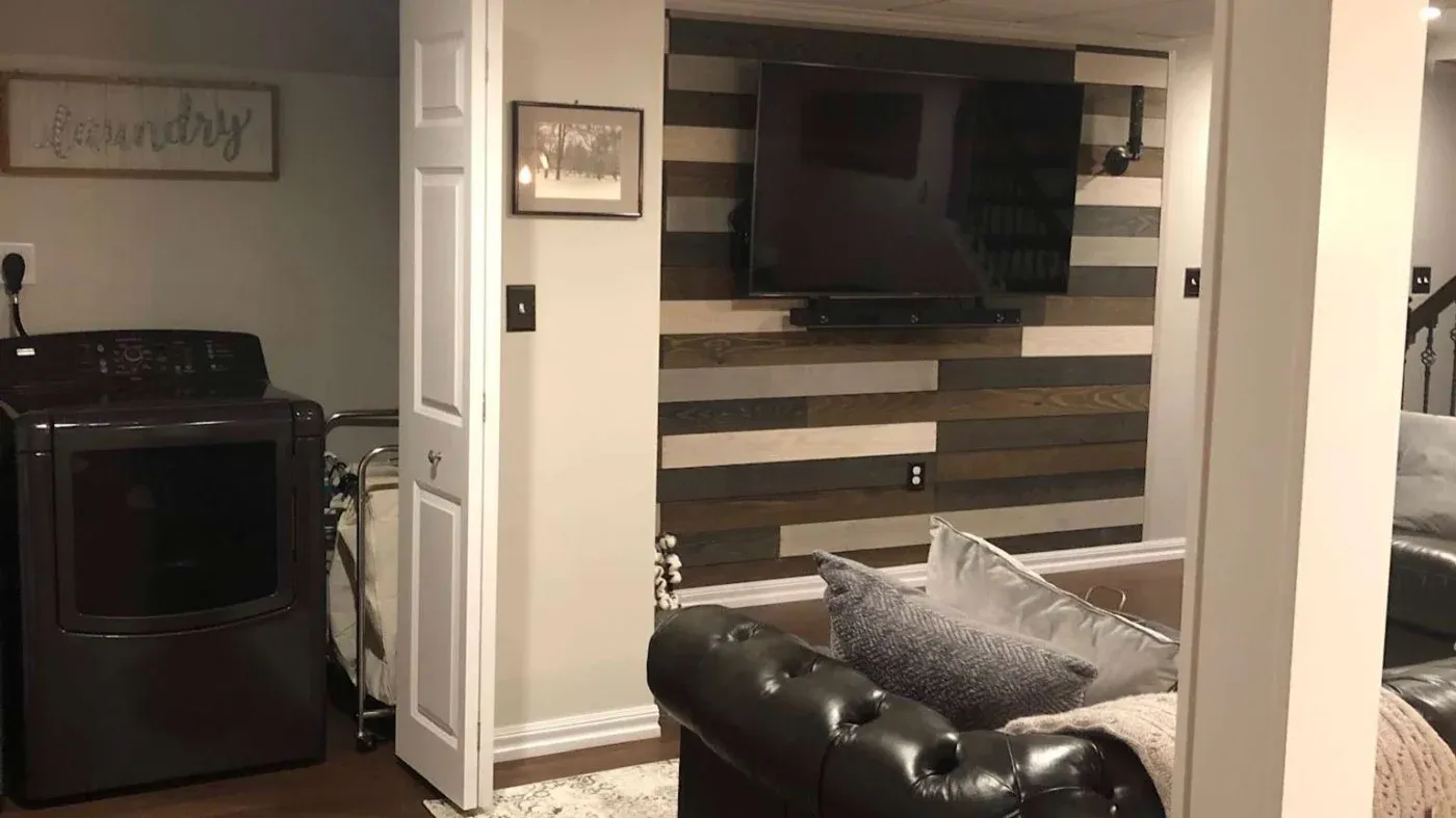 Cozy basement with a flat-screen TV on a wood-planked accent wall. Dark leather sofas, gray pillows, and a laundry area convey warmth and functionality.