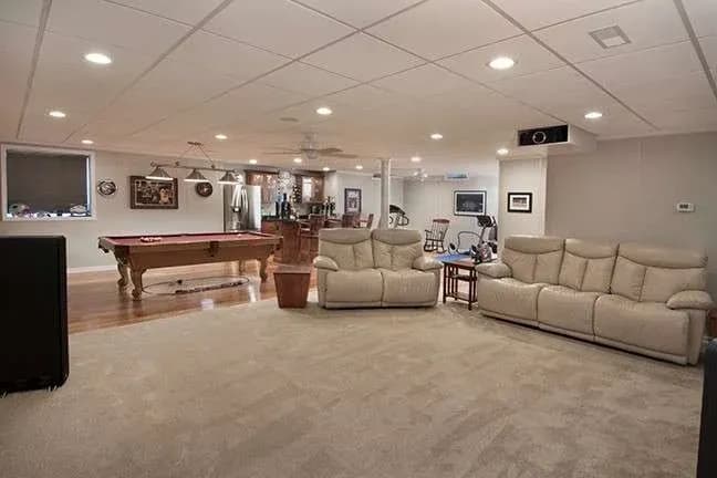 Spacious basement with beige leather sofas on carpet, a pool table on hardwood floor, and exercise equipment. Warm, inviting entertainment area.