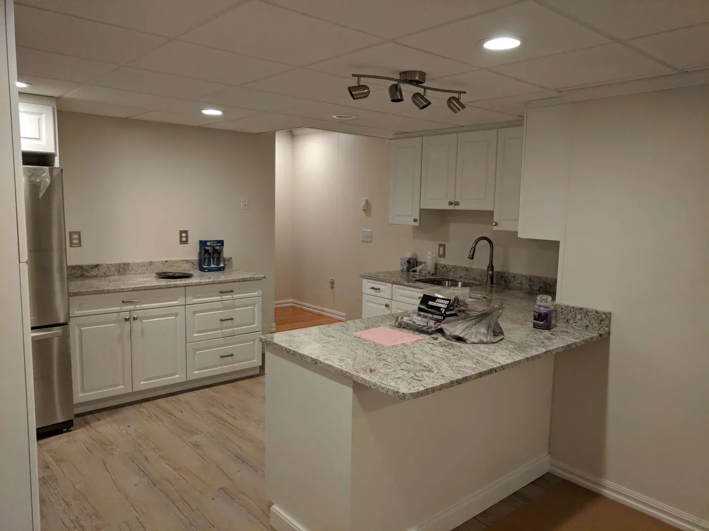 Modern kitchen with white cabinets and granite countertops. Stainless steel fridge on the left, ceiling lights, and light wooden floor create a clean, fresh atmosphere.