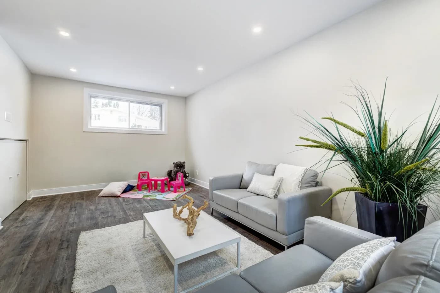 Modern living room with gray sofas, white walls, and dark wood flooring. A small play area with pink chairs, pillows, and toys is in the background.