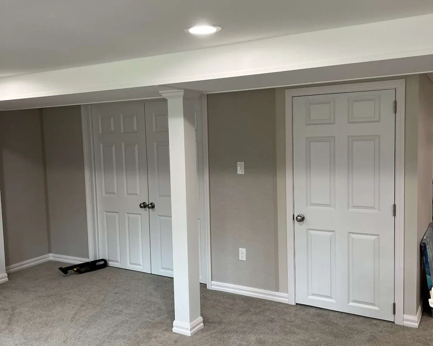 A basement with beige carpet, white walls, and two closed white doors under a low ceiling. A column separates the doors. A light creates a cozy ambiance.