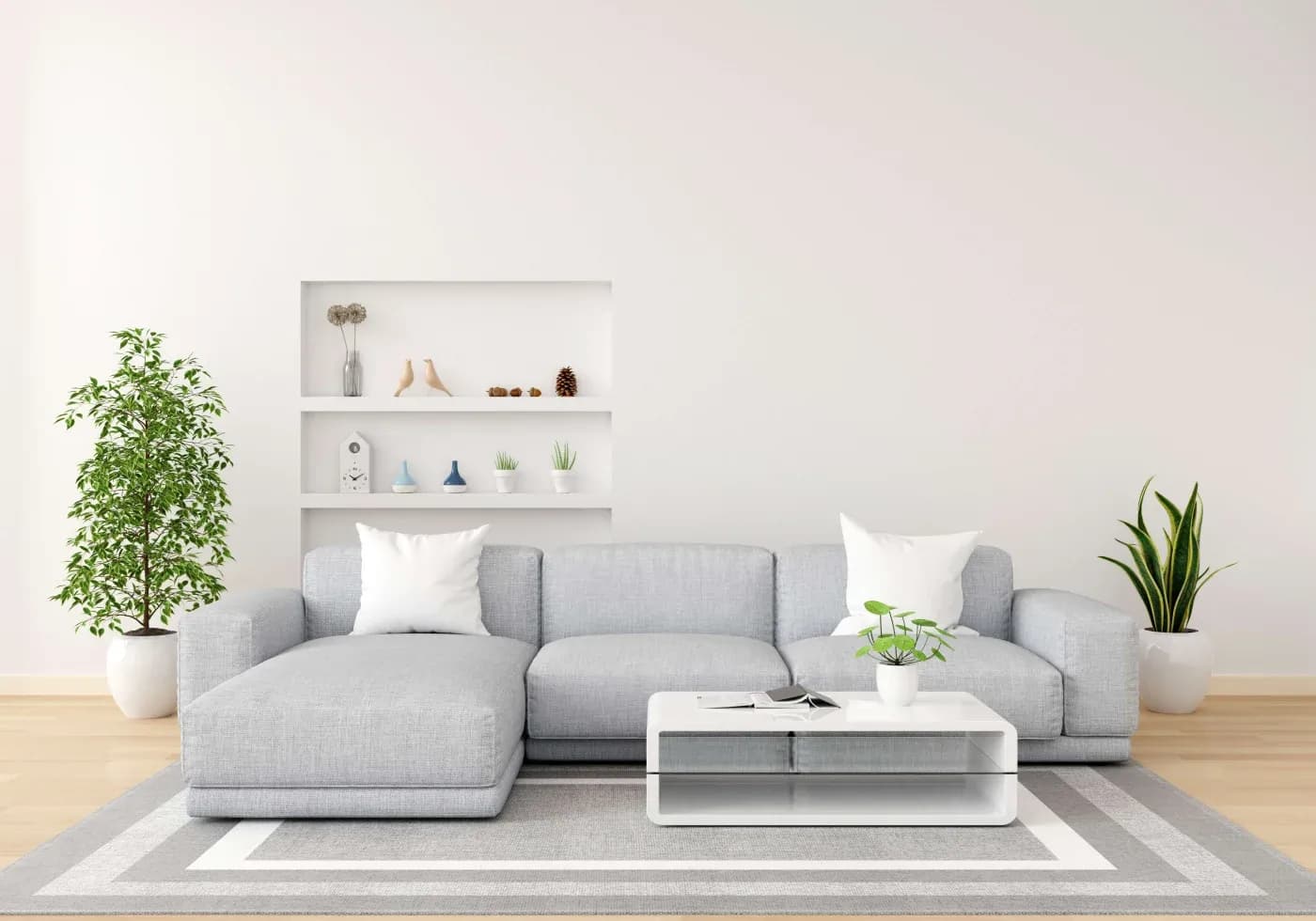A modern living room with a light gray sectional sofa, white pillows, and a white coffee table holding a small plant. Shelves and plants add a minimalist touch.