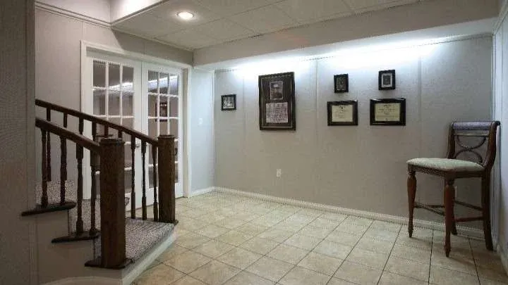A well-lit room with tiled floors and light walls. Stairs with dark wood railing lead to French doors. Framed certificates and a chair adorn the space.