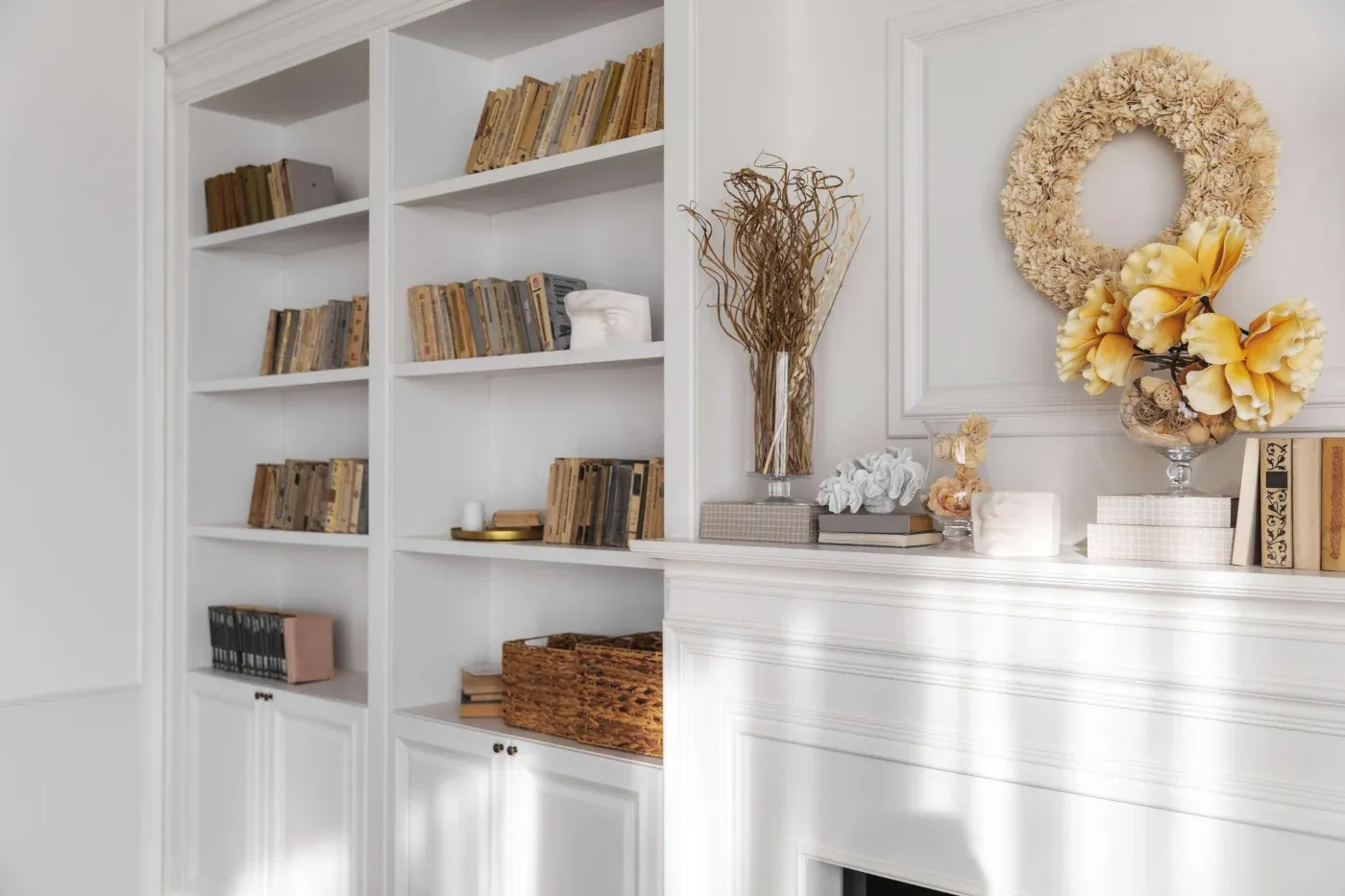 A sunlit, elegant living room features white built-in bookshelves with vintage books and a white mantel adorned with autumn decor and a wreath. The atmosphere is warm and inviting.