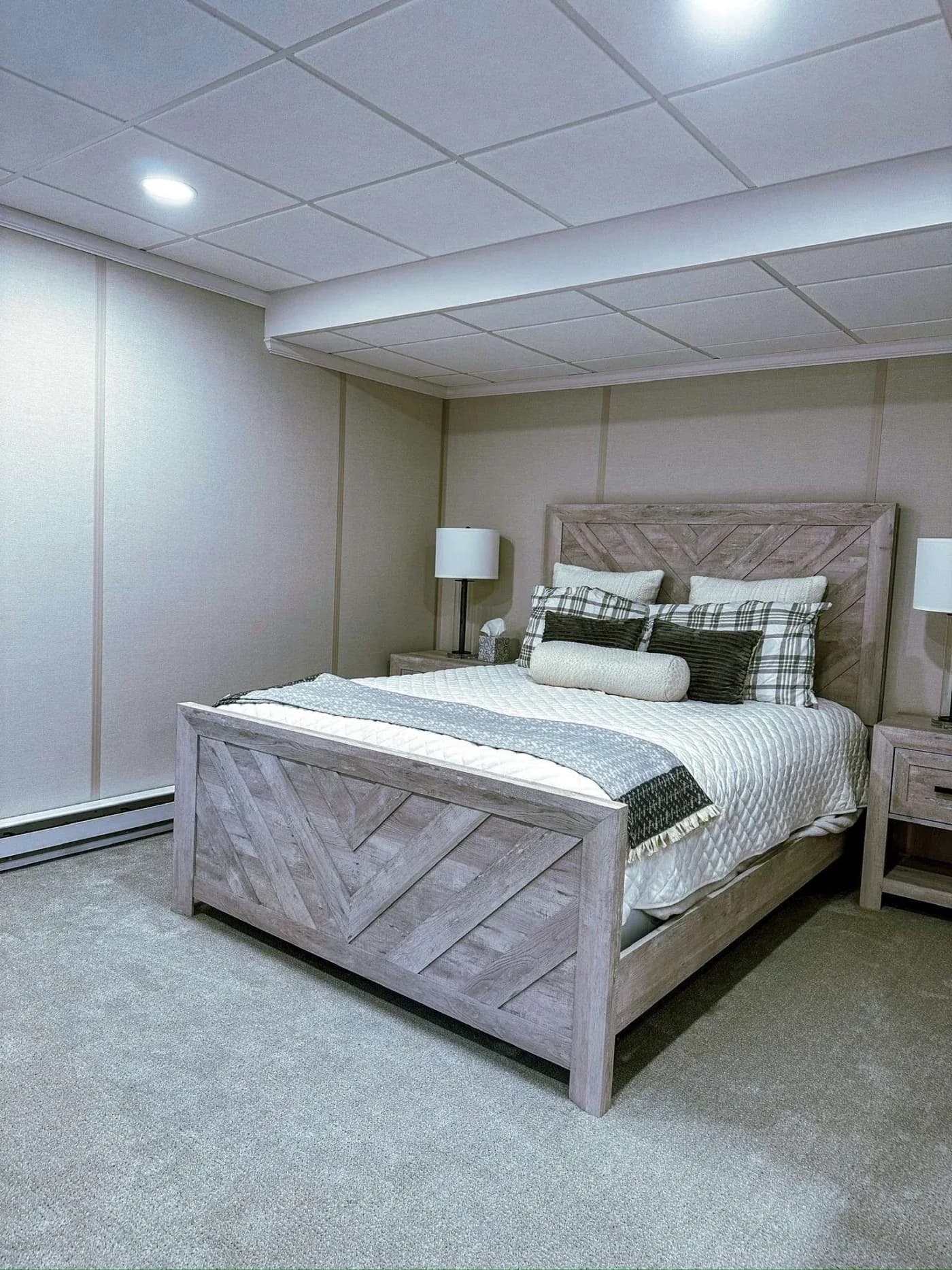 Cozy basement bedroom with a wooden bed, layered with plaid and striped pillows. Soft lighting and neutral walls create a calm, inviting ambiance.