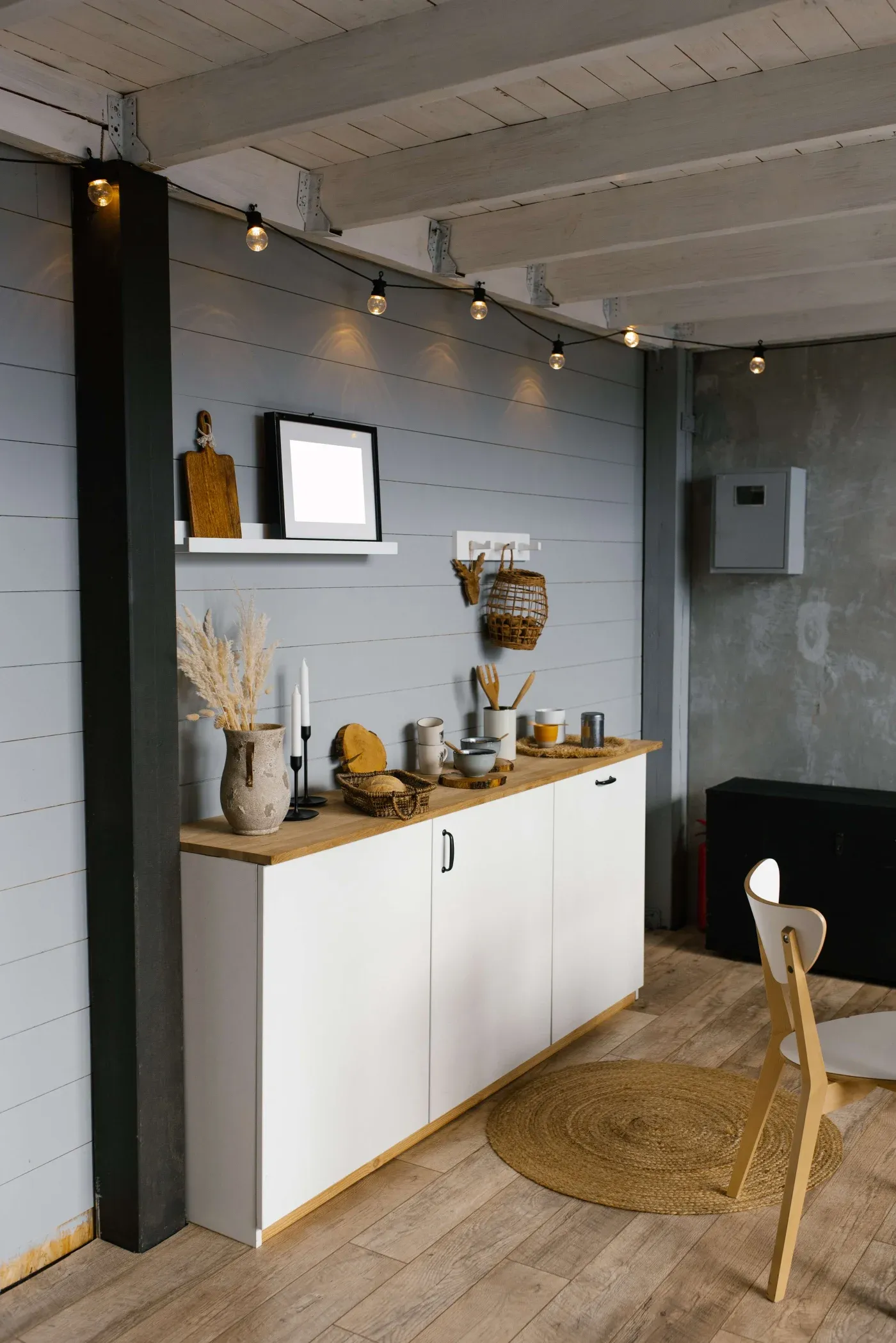 Stylish rustic kitchen area with white cabinets, wood countertop, wicker basket, vases, candles, and hanging string lights, creating a cozy ambiance.